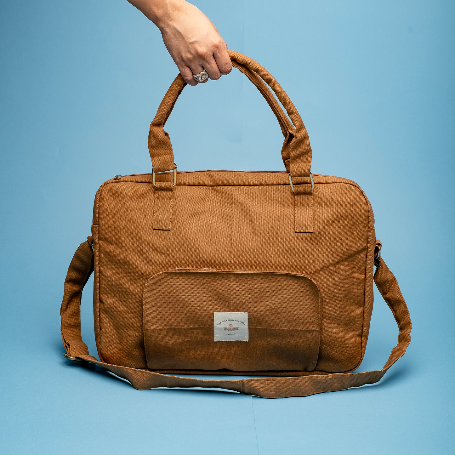 A stylish brown canvas laptop bag with a top handle and detachable shoulder strap. The bag features a front pocket with a small white label and metallic buckle details on the straps, displayed against a blue background.