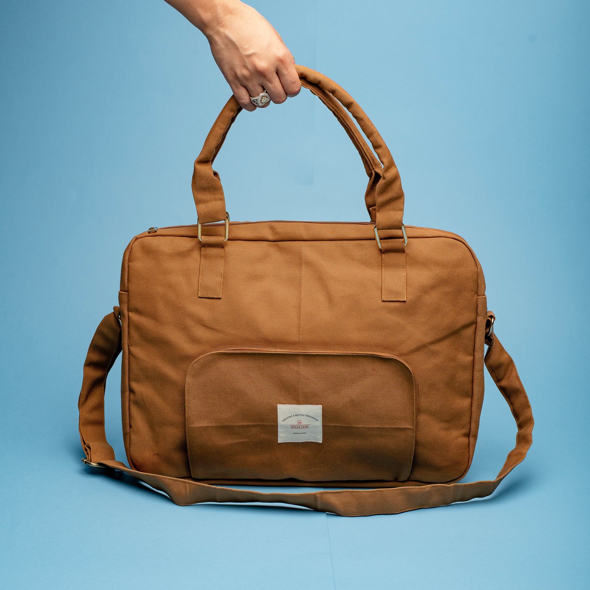 A stylish brown canvas laptop bag with a top handle and detachable shoulder strap. The bag features a front pocket with a small white label and metallic buckle details on the straps, displayed against a blue background.