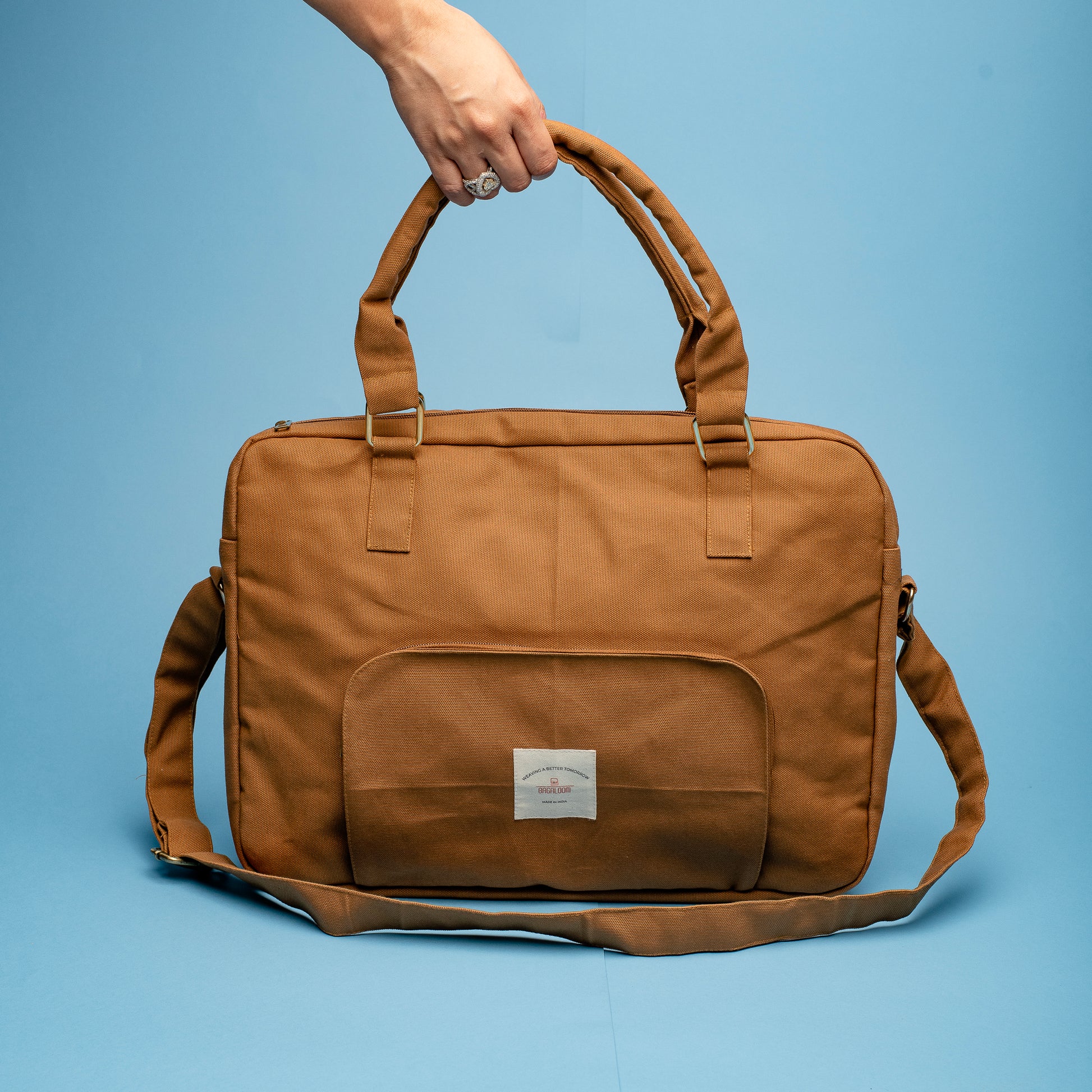 A stylish brown canvas laptop bag with a top handle and detachable shoulder strap. The bag features a front pocket with a small white label and metallic buckle details on the straps, displayed against a blue background.

