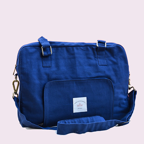 A stylish blue canvas laptop bag with a top handle and detachable shoulder strap. The bag features a front pocket with a small white label and metallic buckle details on the straps, displayed against a pink background.

